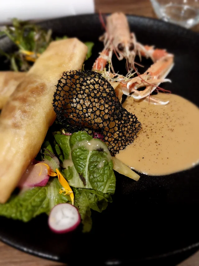 Assiette noire avec dedans : une langoustine, feuilles vertes, sauce, décor noir et une crêpe