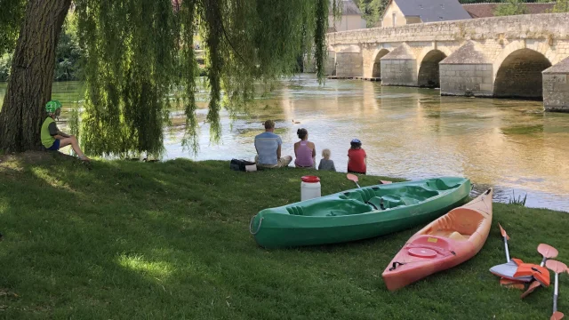 Canoé en bord de rivière avec une famille au pied d'un arbre avec vélos au premier plan, pont en pierre et forteresse en arrière plan