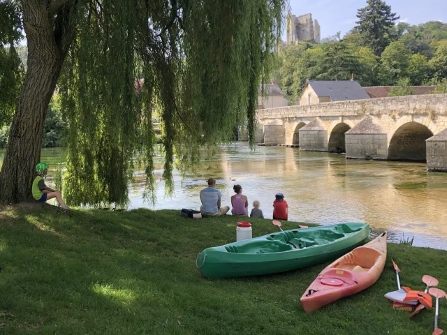 Canoé en bord de rivière avec une famille au pied d'un arbre avec vélos au premier plan, pont en pierre et forteresse en arrière plan